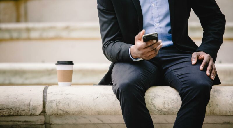 crop businessman using smartphone while resting on bench with takeaway coffee